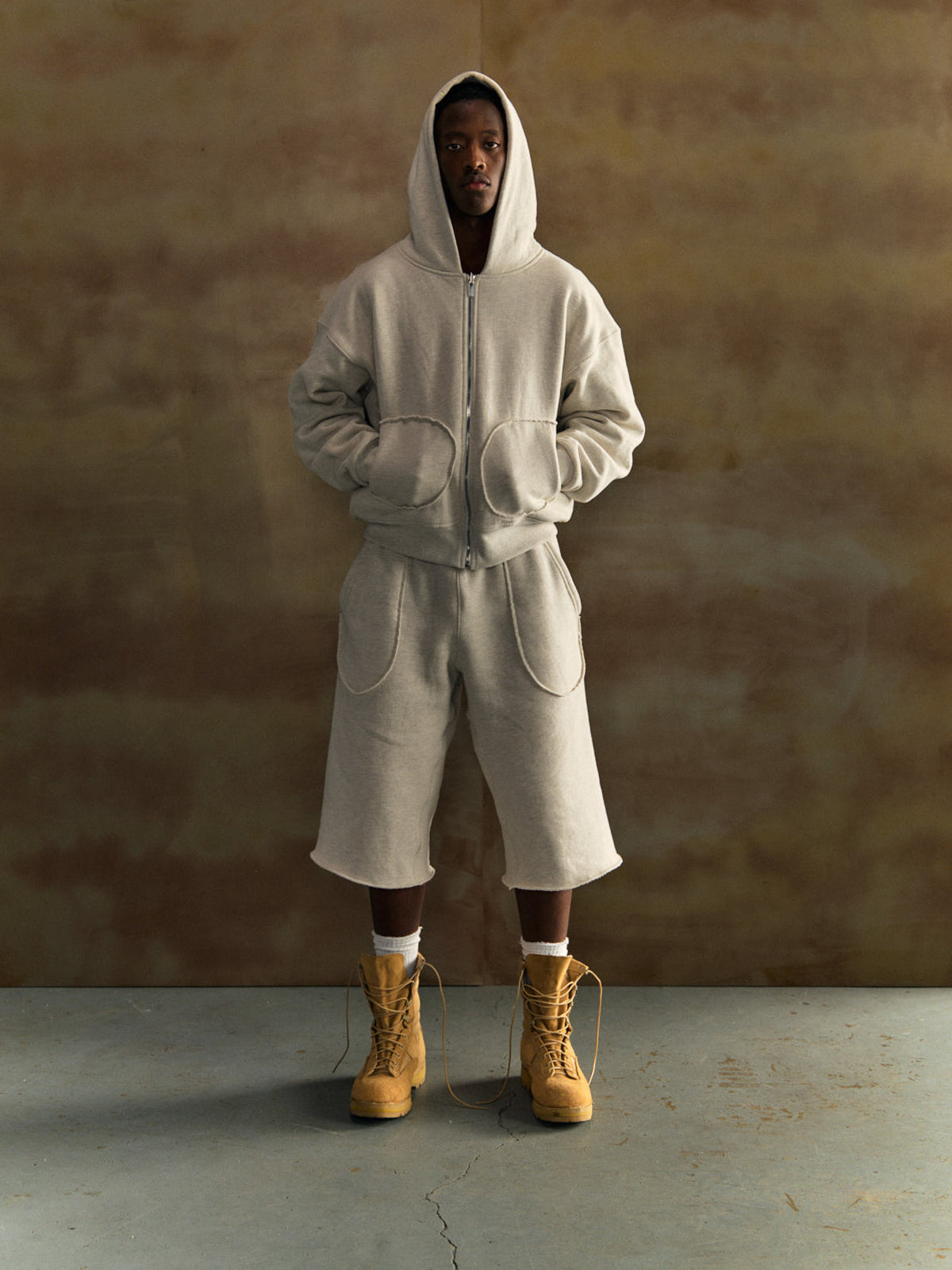 Person wearing a gray tracksuit and yellow boots against a brown background