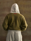 Person wearing a green hoodie and gray pants with a hood, standing against a brown background
