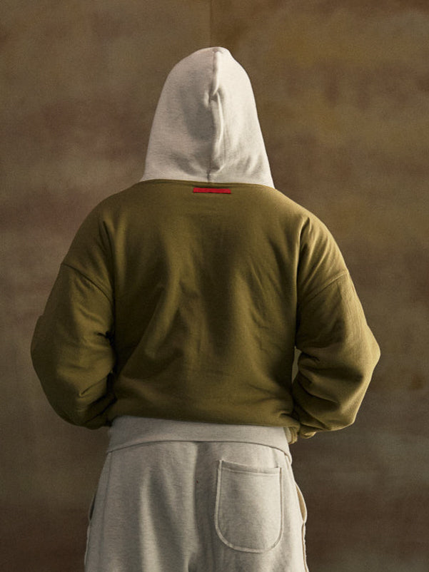 Person wearing a green hoodie and gray pants with a hood, standing against a brown background