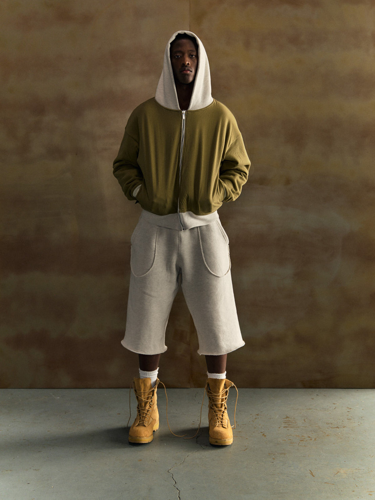 Person wearing a green hoodie, gray pants, and yellow boots against a brown background