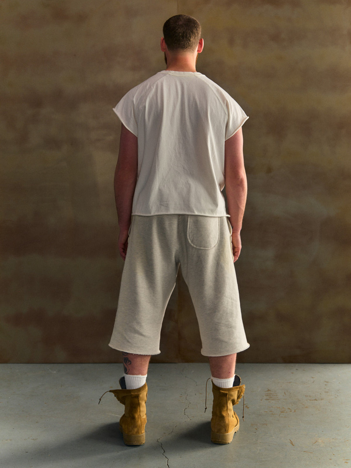 Person wearing a white t-shirt and beige shorts with brown boots against a textured wall.